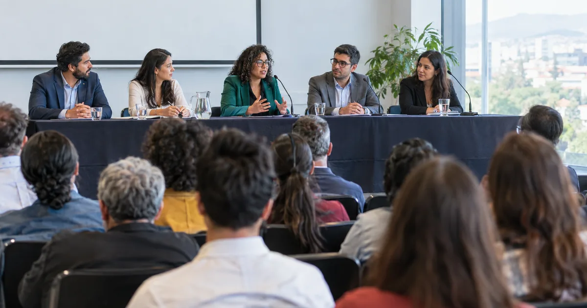 Imagen generada con IA: panel de ponentes en una mesa larga frente a un público en sala (escena ilustrativa con personas ficticias)
